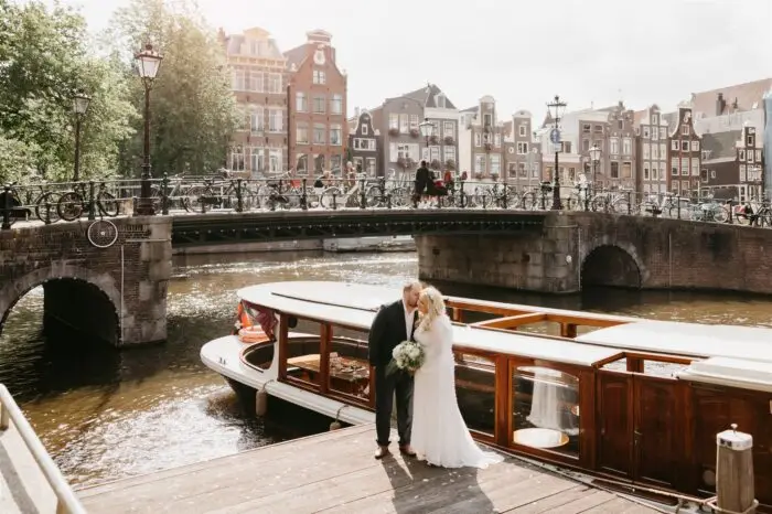 Couple standing by a canal boat in Amsterdam with a historic bridge and canal houses in the background