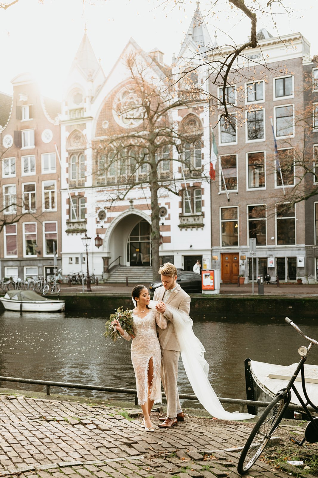 Bridal couple standing by the canal with Keizersgrachtkerk in the background during their photoshoot in Amsterdam