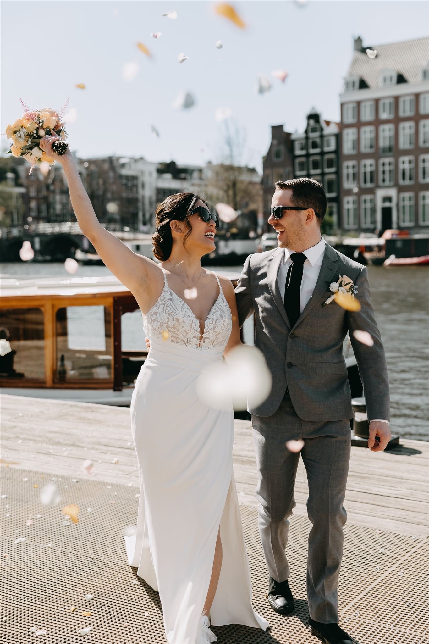Happy bridal couple walking on a canal pier after their Amsterdam elopement ceremony, bride holding bouquet up