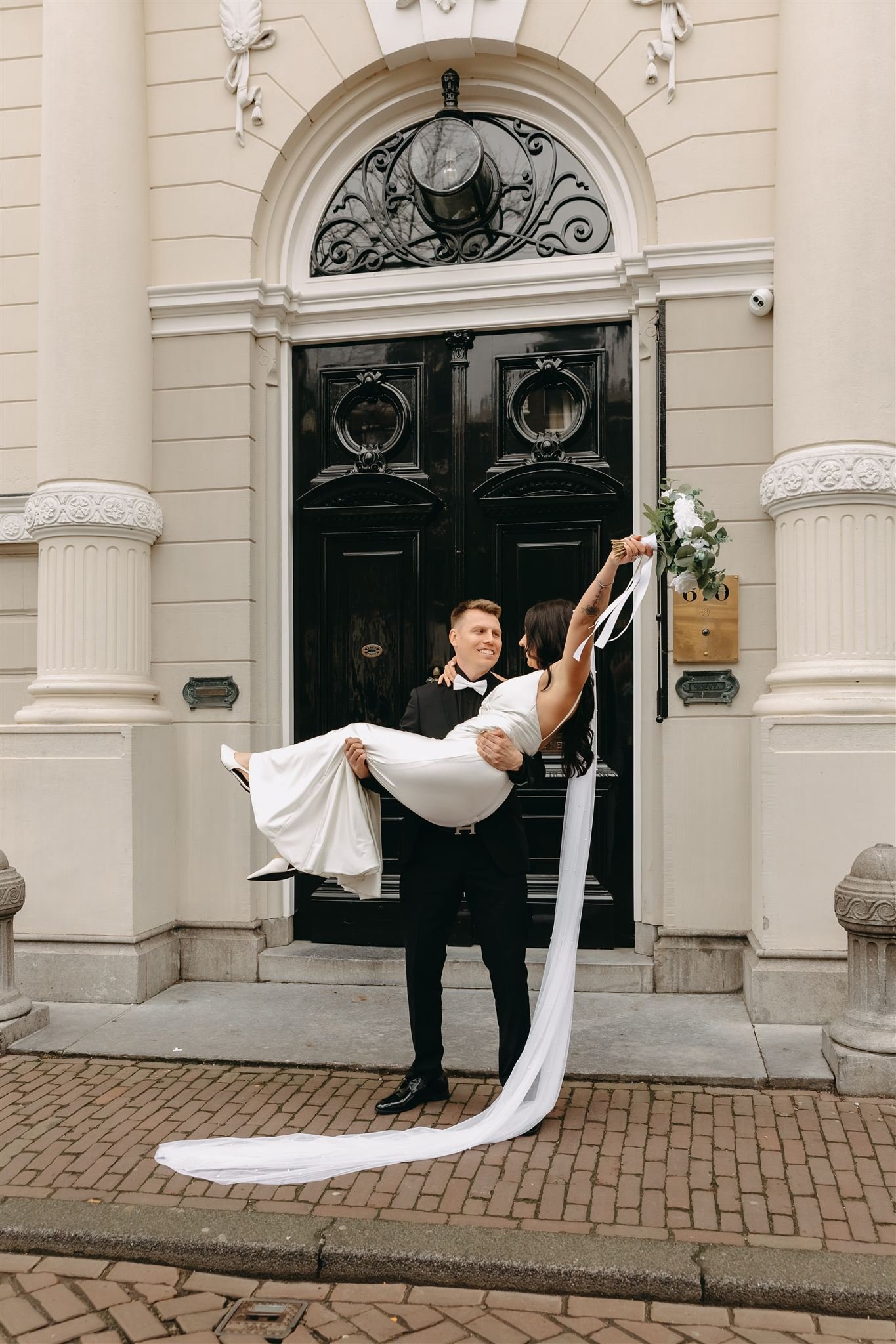 Groom carrying his bride in front of a historic house in Amsterdam, both smiling and joyful
