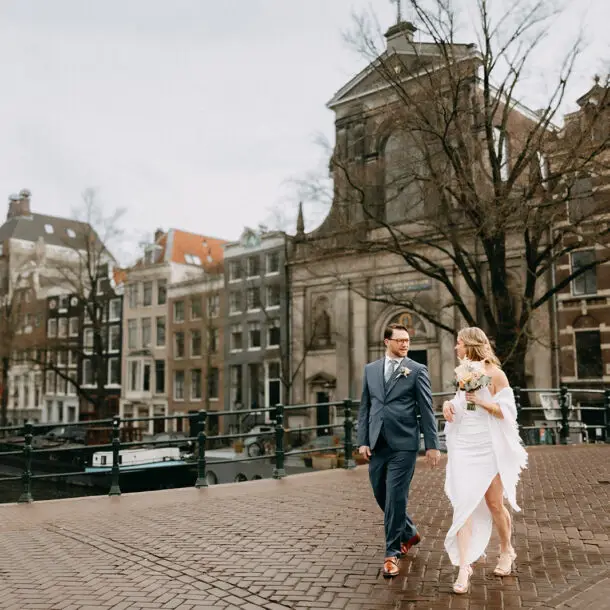 Couple walking on a canal bridge during their elopement in Amsterdam with historic buildings and church in the background