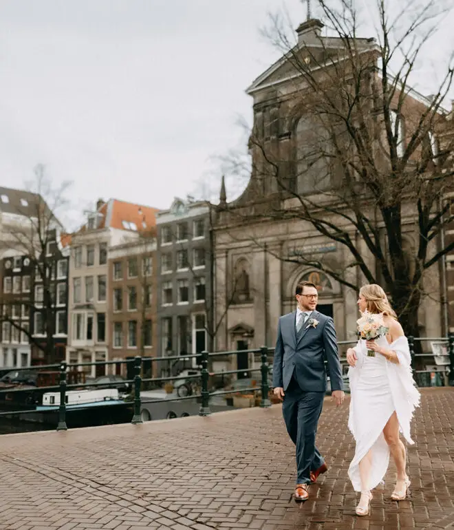 Couple walking on a canal bridge during their elopement in Amsterdam with historic buildings and church in the background