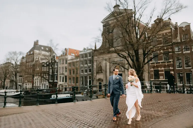 Couple walking on a canal bridge during their elopement in Amsterdam with historic buildings and church in the background