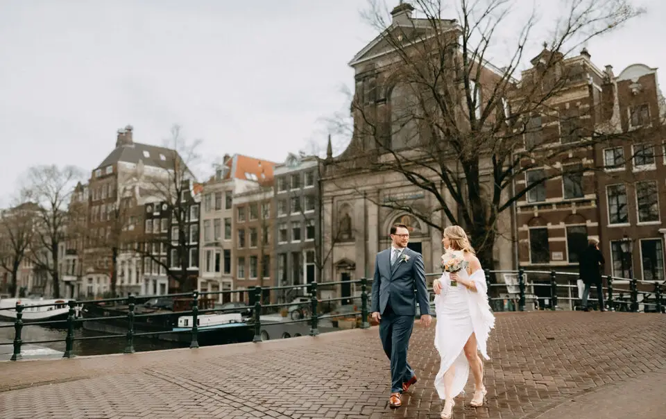 Couple walking on a canal bridge during their elopement in Amsterdam with historic buildings and church in the background