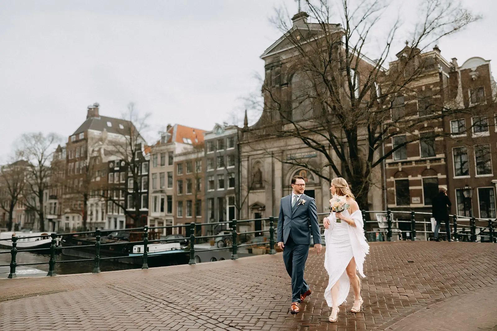 Couple walking on a canal bridge during their elopement in Amsterdam with historic buildings and church in the background