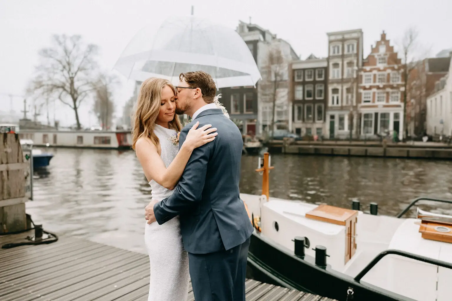 Kristin and Logan embracing under a transparent umbrella beside their private canal boat in Amsterdam