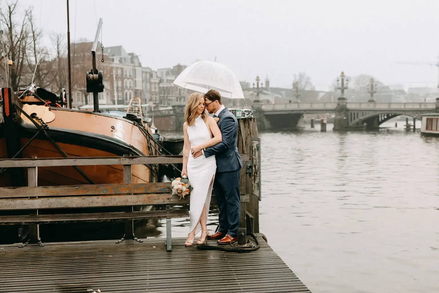 Kristin and Logan embracing under a transparent umbrella beside the water during their Amsterdam wedding photoshoot
