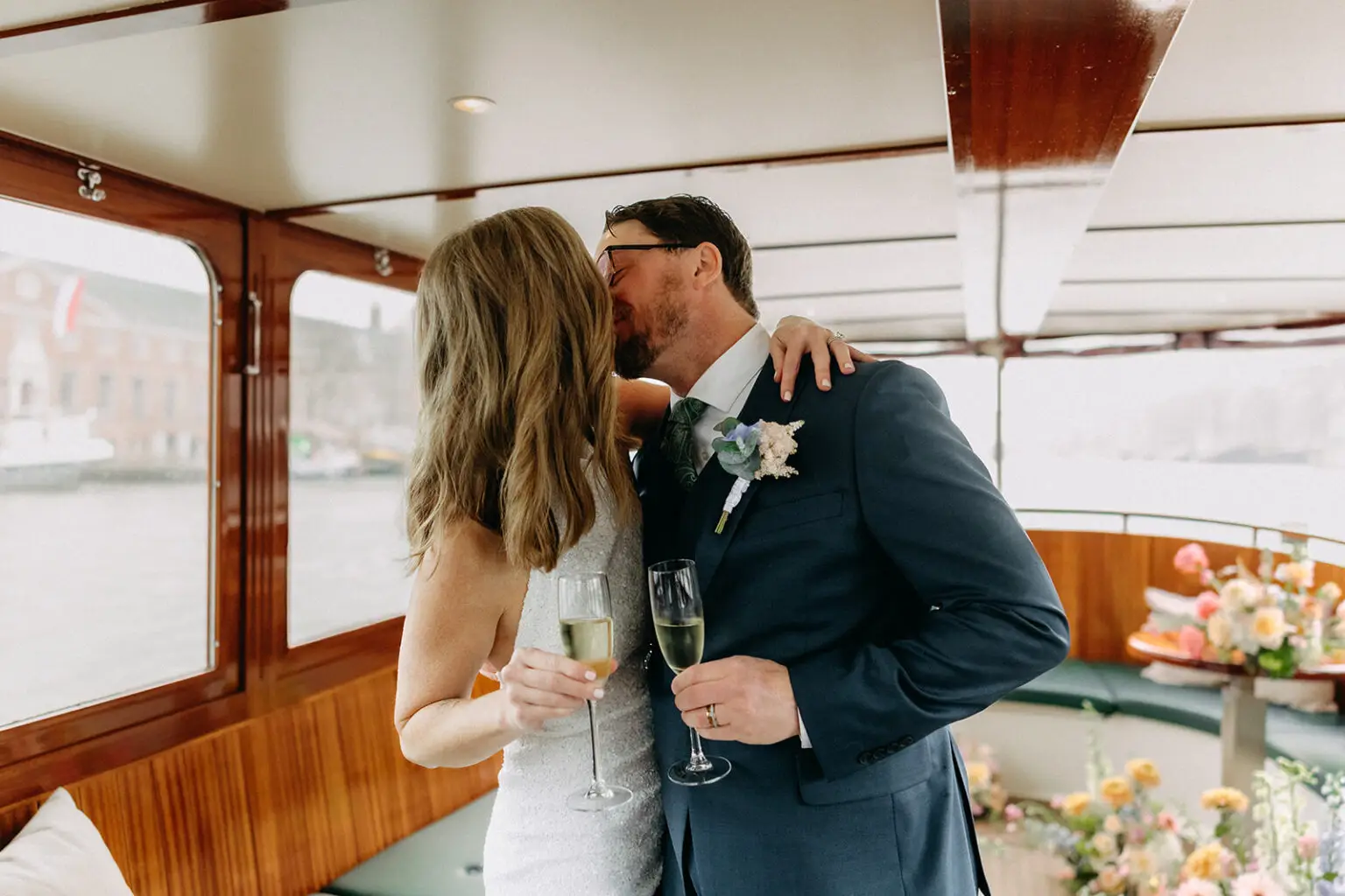 Kristin and Logan sharing a kiss while holding champagne glasses aboard their private canal boat in Amsterdam