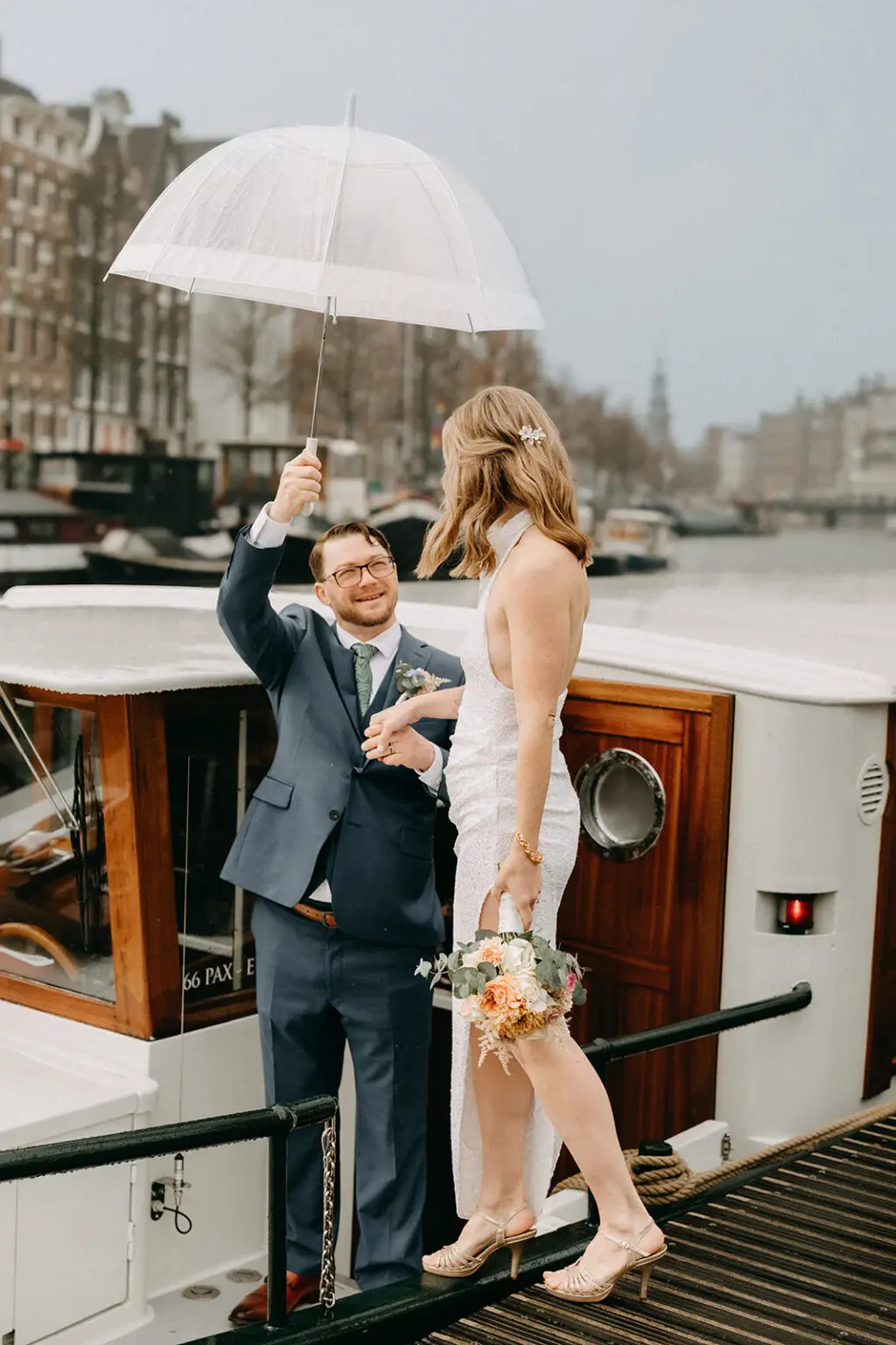 Groom holding a transparent umbrella for his bride during a rainy canal boat wedding in Amsterdam