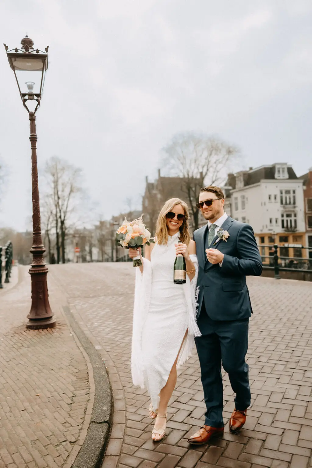 Kristin and Logan walking across an Amsterdam canal bridge with champagne and wedding bouquet after their ceremony