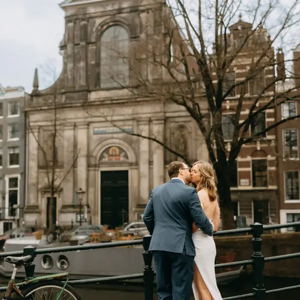 Bride and groom embracing beside an Amsterdam canal with a historic church and canal boat during a wedding photoshoot in Amsterdam