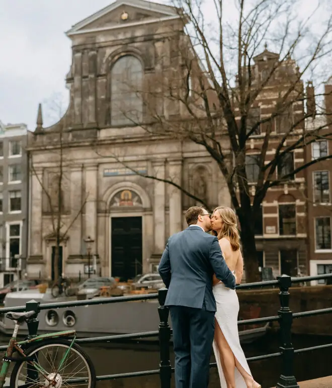 Kristin and Logan embracing beside an Amsterdam canal with a historic church and canal boat in the background