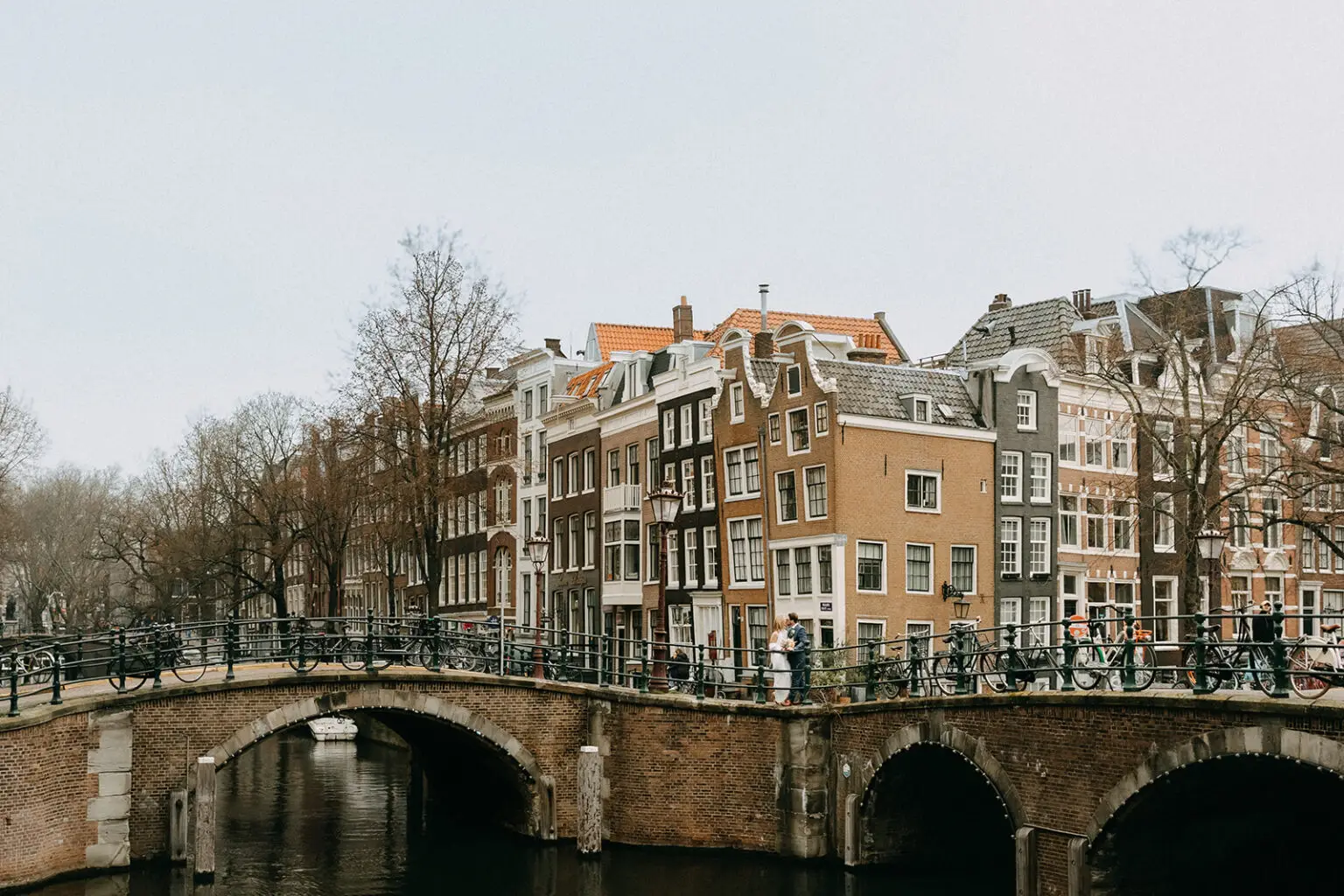 Kristin and Logan standing on a historic canal bridge surrounded by Amsterdam canal houses during their wedding photoshoot