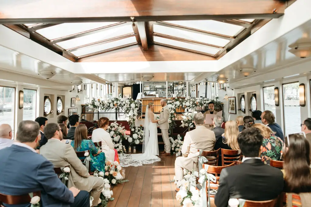 Bride and groom exchanging vows during a symbolic ceremony aboard Het Wapen van Amsterdam surrounded by elegant white floral arrangements