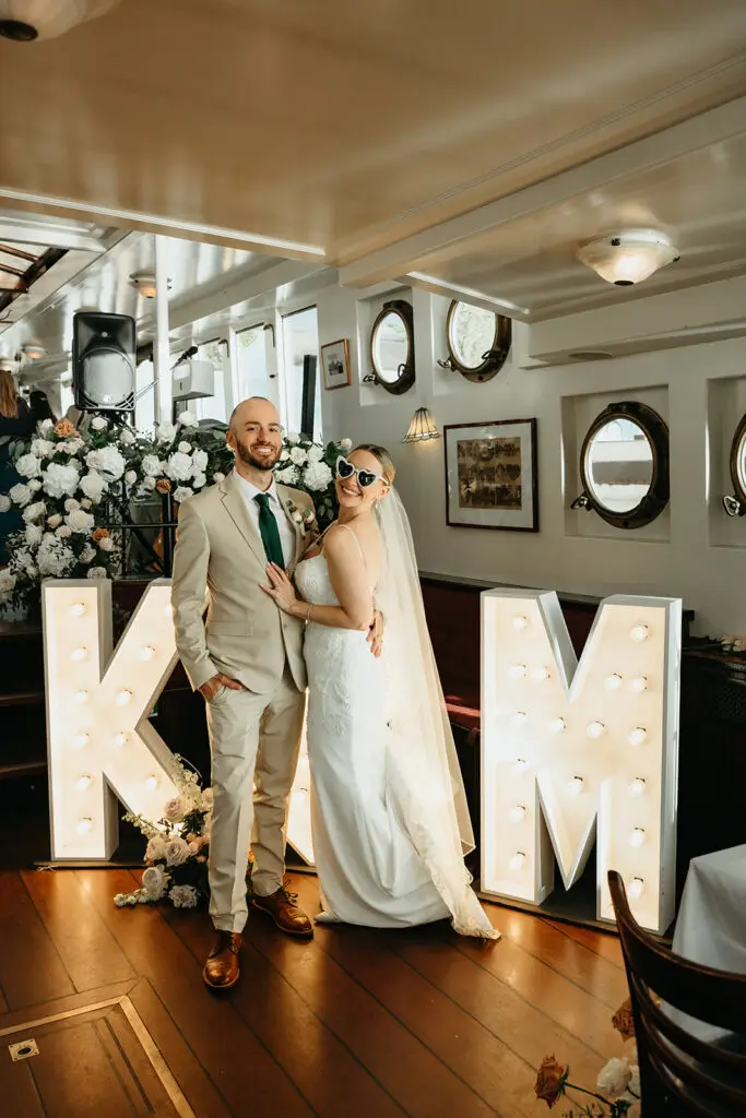 Bride and groom posing beside illuminated initials during their Amsterdam destination wedding reception aboard a canal boat